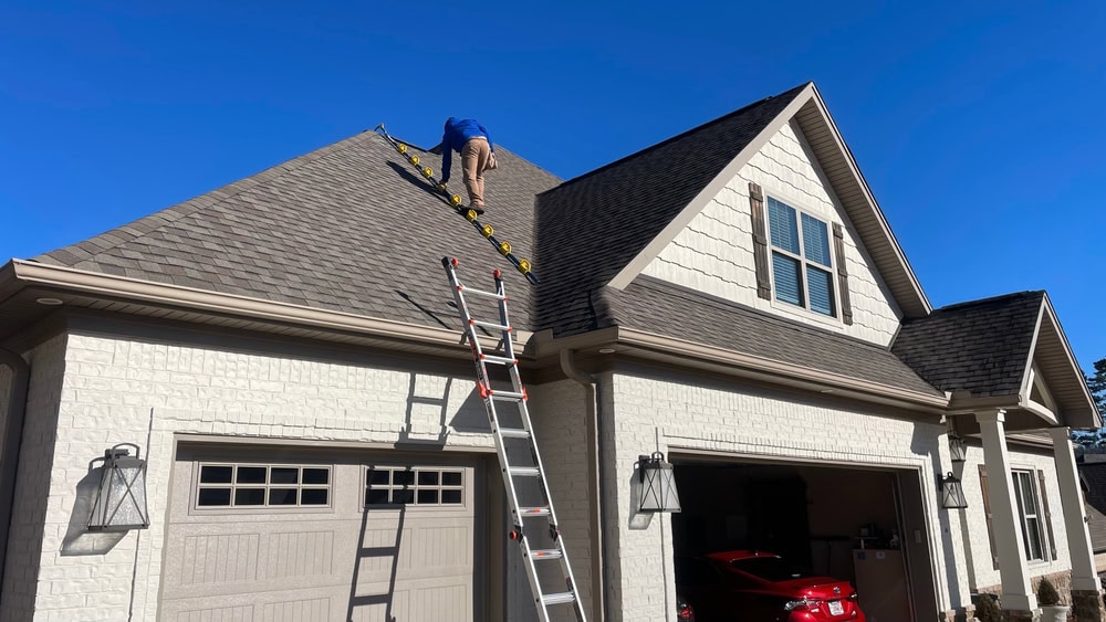 A person wearing a blue shirt is using a ladder to climb onto the steep roof of a light-colored house with two garages; a red car is parked inside one garage under a clear blue sky.