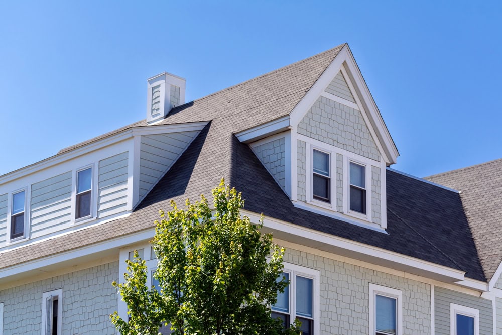 A light gray house with white trim, multiple windows, and an angled roof showcases quality residential roofing Broward County, FL. A leafy green tree partially obscures the lower part of the building under a clear blue sky.