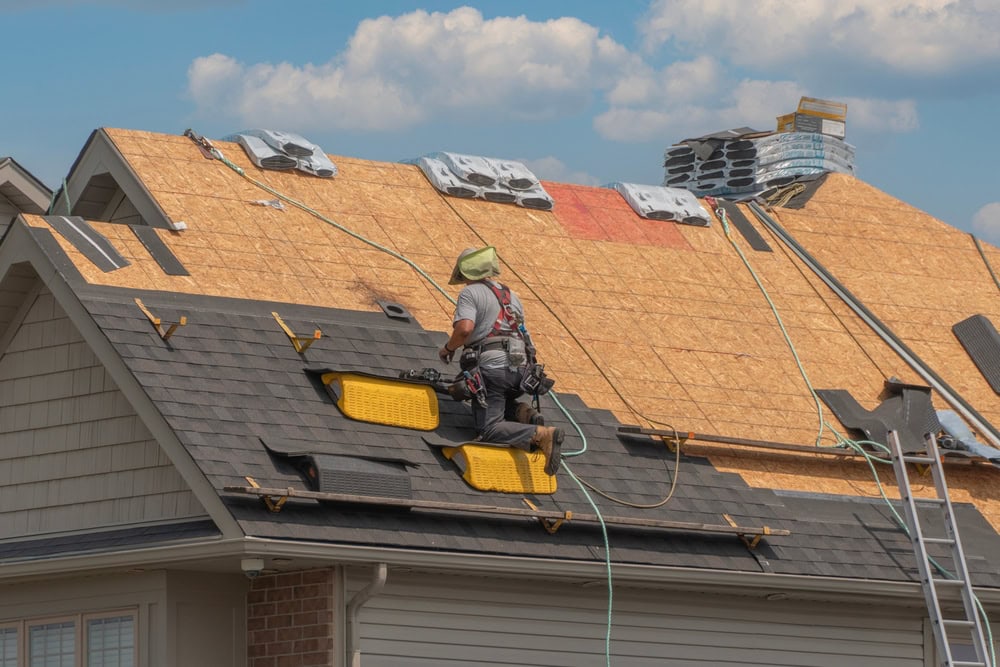 A construction worker in safety gear installs shingles on the roof of a house under a partly cloudy sky. Roofing materials and tools are visible on the unfinished wooden roof.
