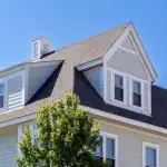 A light gray house with white trim and multiple gabled roofs sits under a clear blue sky. Several windows are visible, and a green leafy tree partially obscures the lower part of the house.
