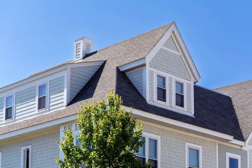 A light gray house with white trim and multiple gabled roofs sits under a clear blue sky. Several windows are visible, and a green leafy tree partially obscures the lower part of the house.