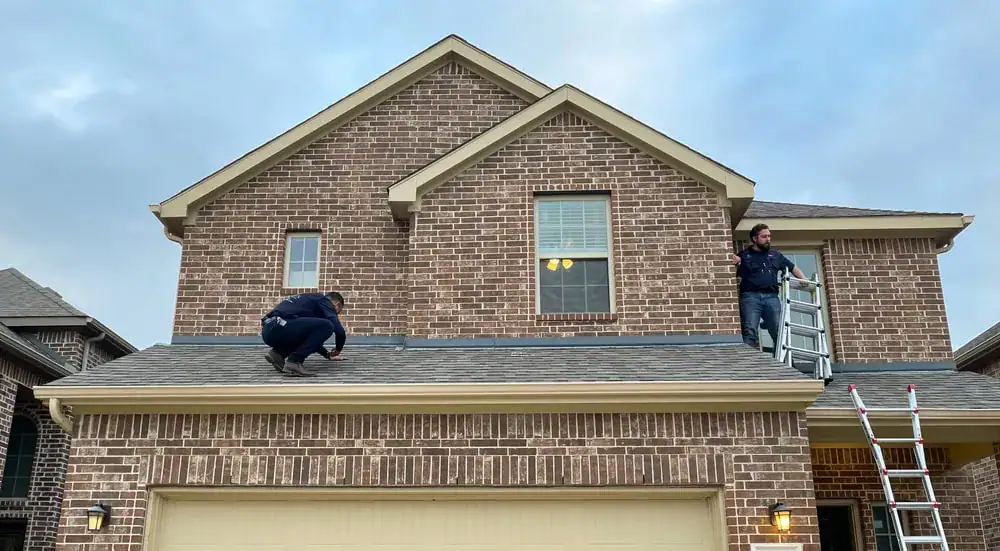 Two people are inspecting the roof of a two-story brick house. One person squats near the edge, while the other stands near a ladder, holding it and looking toward the crouched person. The sky is cloudy.
